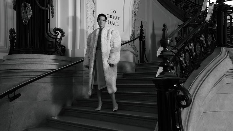 Black and white shot of a woman in a fur coat on a staircase, with a marble railing and architectural details in the background.