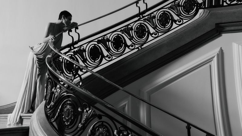 Monochrome photo of a woman in gown leaning on decorative staircase indoors. The image highlights architectural and fashion elements.