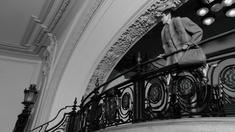 Woman in fur coat and dress holding a purse on an ornate wrought iron staircase, in a classic interior with architectural details.