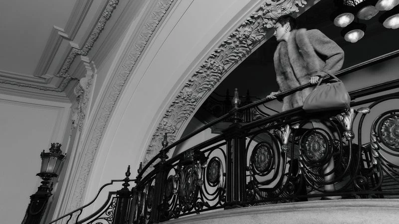 Woman in fur coat and dress holding a purse on an ornate wrought iron staircase, in a classic interior with architectural details.