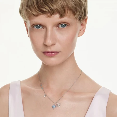 Close-up of a light-skinned woman with short blonde hair, wearing a delicate silver necklace with blue and white charms on a white backdrop.