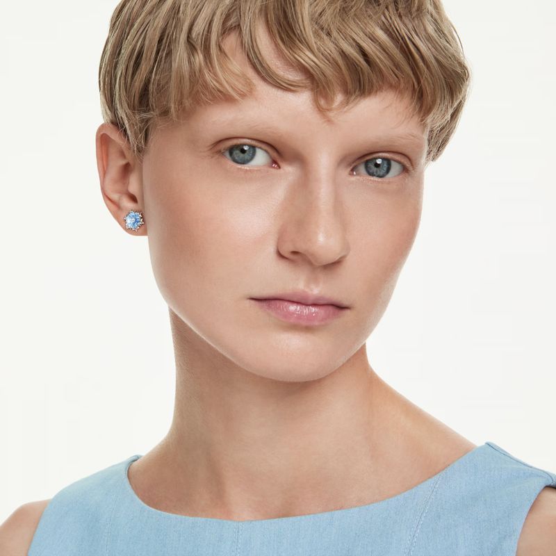 Close-up studio shot of a fair-skinned young woman with blue eyes and short blonde hair, wearing light blue earrings and a matching top.