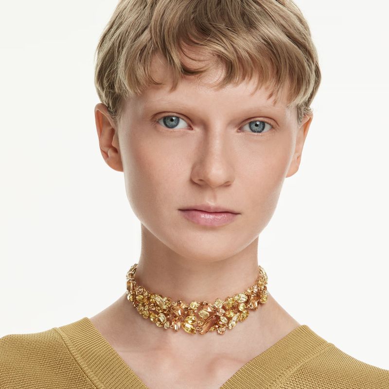 Close-up studio shot of a blond woman wearing a gold choker necklace and a gold top against a white background in portrait orientation.
