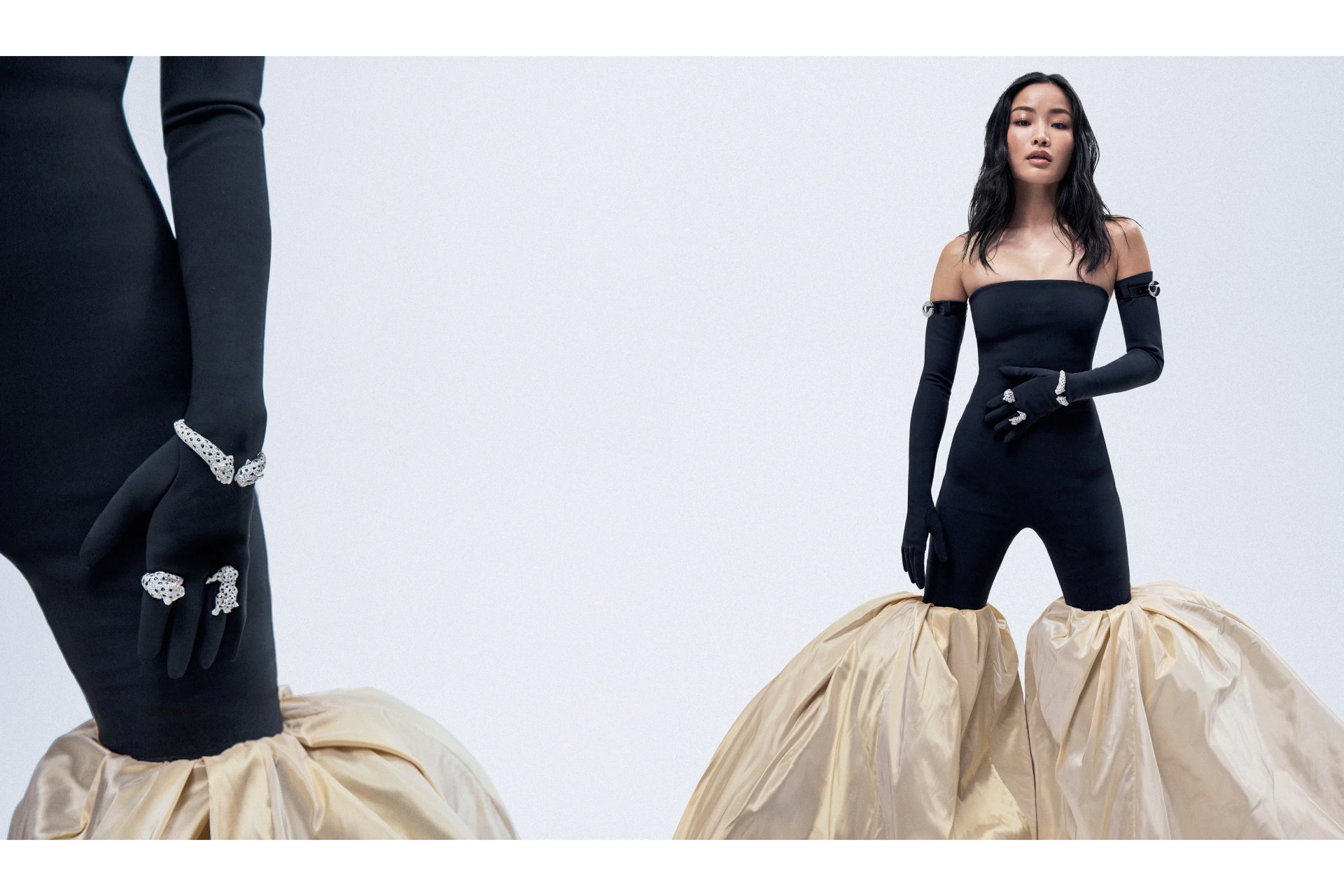 A slender woman poses in a studio wearing a black jumpsuit, long black gloves, bracelets, and rings with a tan ruffled lower-skirt.