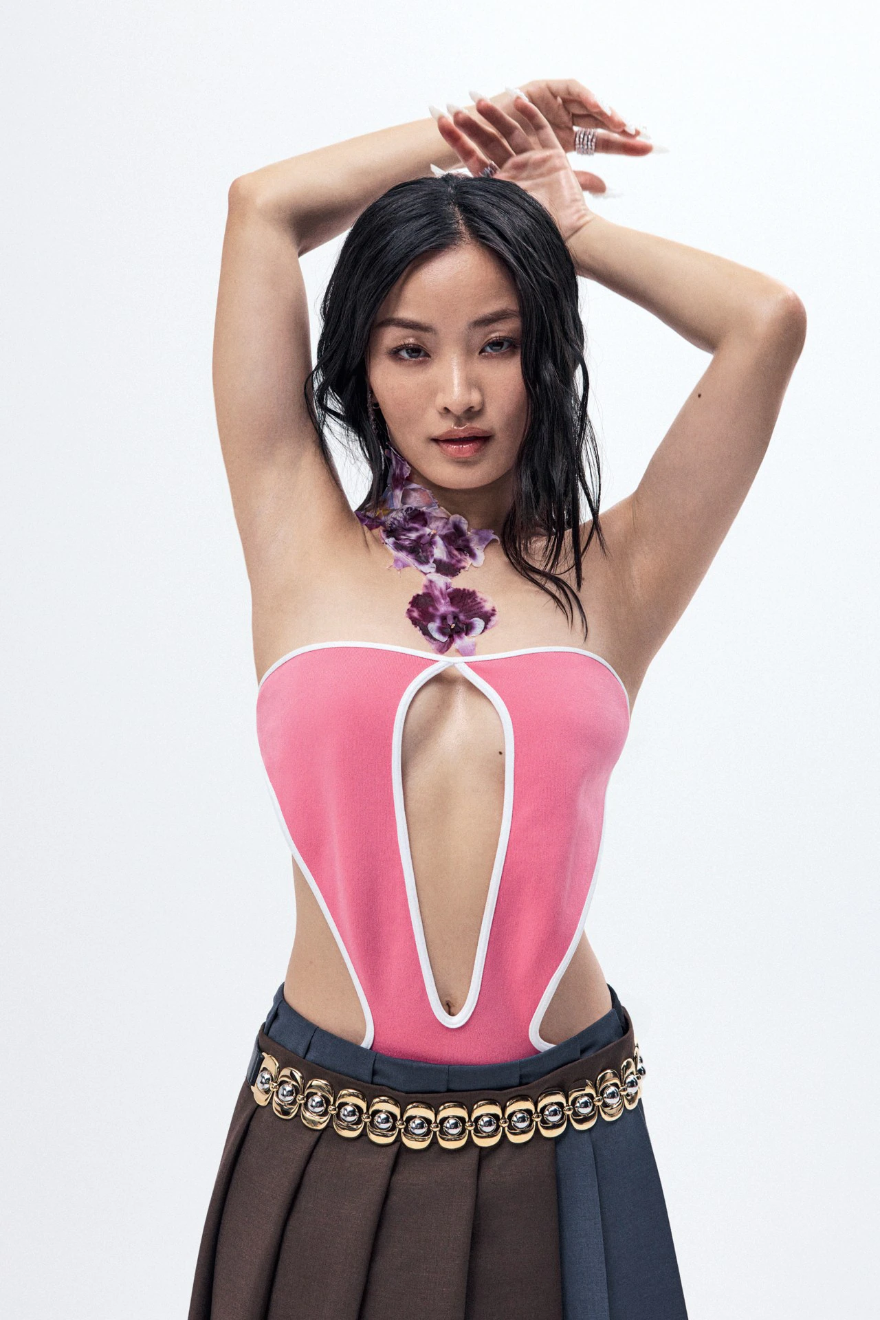 Anna Sawai wearing pink cutout top and pleated skirt with metallic-studded belt against plain white background