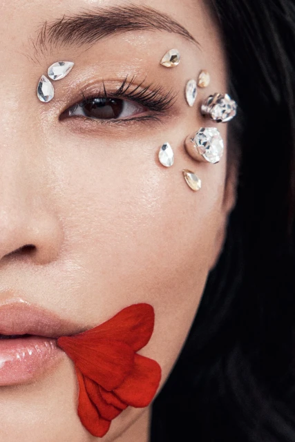 Close-up portrait of an Asian woman with jewel embellishments around her eye and a red flower petal near her lips.
