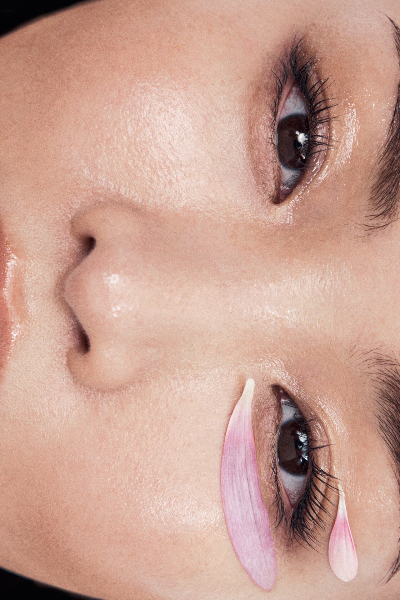 Close-up portrait of an adult woman's face with flower petals near her eye, pale skin, and dramatic eye makeup.