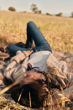 Woman in jeans lies on a blanket in a sunny field surrounded by tall grass, enjoying a peaceful moment in the countryside.