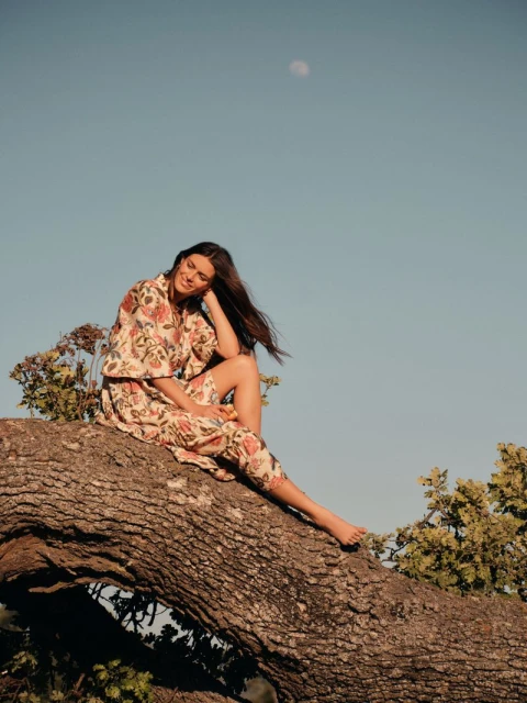 A brunette adult female poses in a floral dress on a tree branch against a clear sky and moon, embodying a natural and relaxed fashion aesthetic.