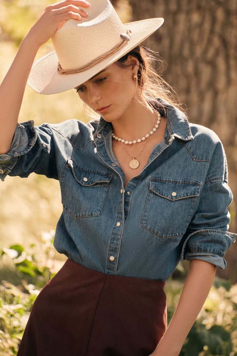 Portrait of a woman in a denim shirt and straw hat, wearing pearl jewelry, posing outdoors with a blurred nature background.