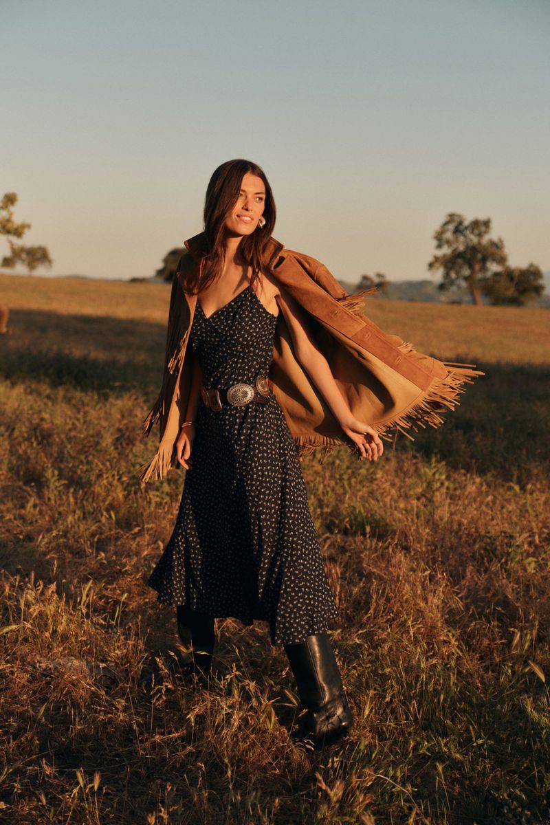 Woman in a printed dress with a fringed suede jacket, standing in a field at golden hour, wearing boots and a concho belt.