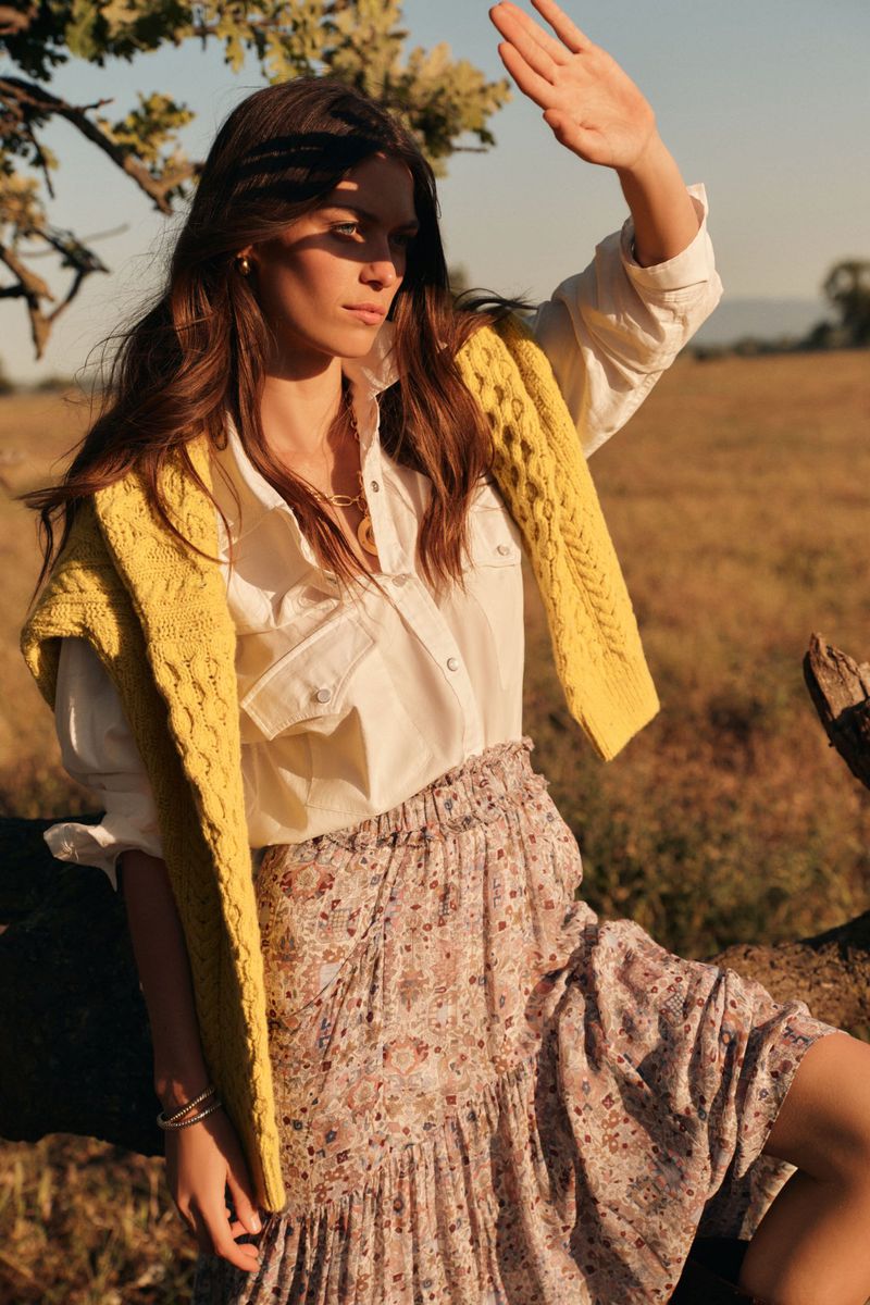A young woman in outdoor golden-hour light shields her face, wearing a white shirt, patterned skirt, and yellow sweater over her shoulders.