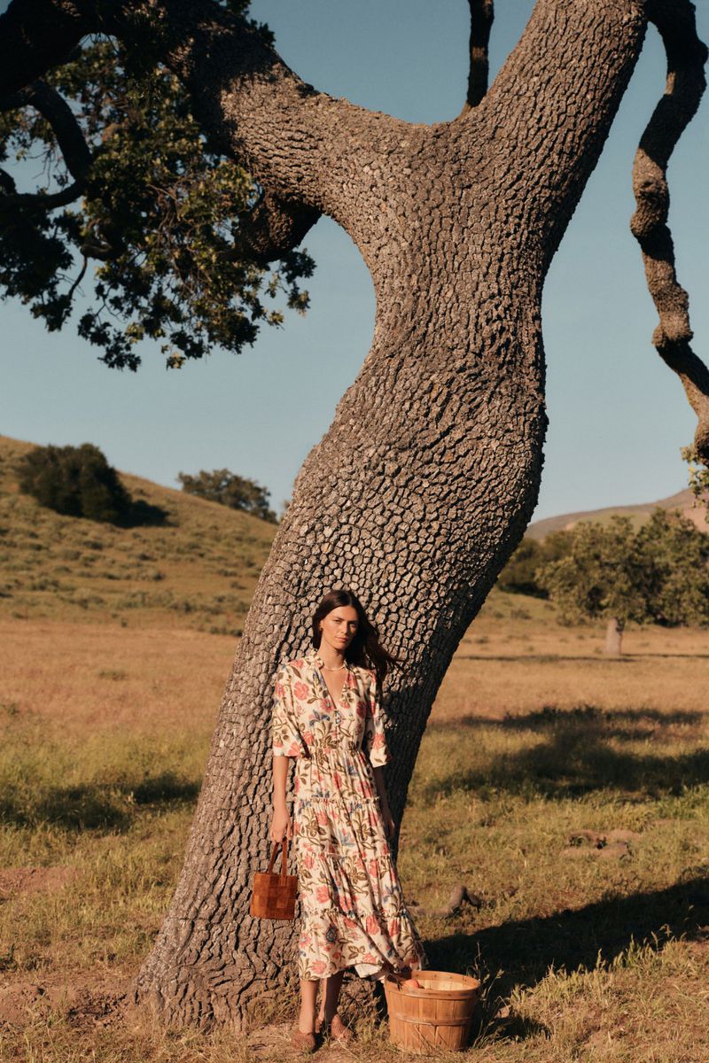 Adult woman poses with brown leather purse in front of large textured tree with rural landscape in background during daytime.