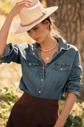 Model wearing a straw cowboy hat, denim shirt, and pearl necklace adjusts her hat outdoors in sunlight.