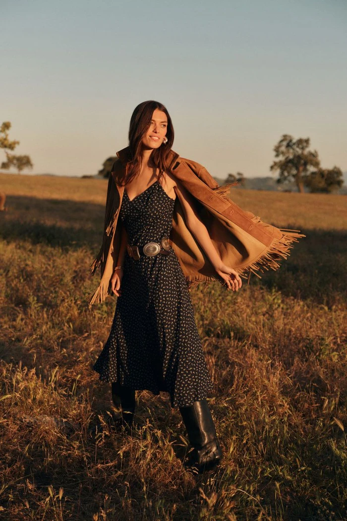 Model George Midgley wearing a polka dot dress and fringed suede jacket in a sunlit grassy field for Tuckernuck.