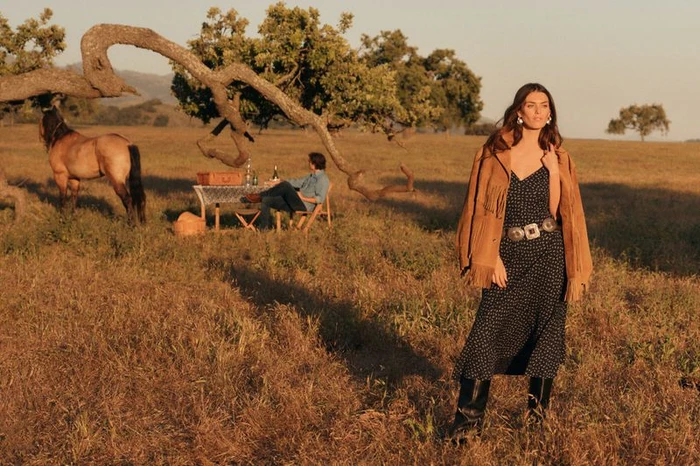 Model wearing a fringed suede jacket and polka dot dress standing in a grassy meadow under golden hour sunlight.