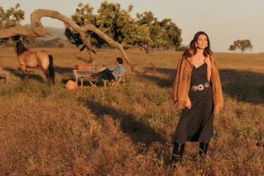 Model wearing a fringed suede jacket and polka dot dress standing in a grassy meadow under golden hour sunlight.