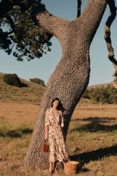 Model wearing a floral dress standing beside a large oak tree trunk in an outdoor grassland setting for Tuckernuck.