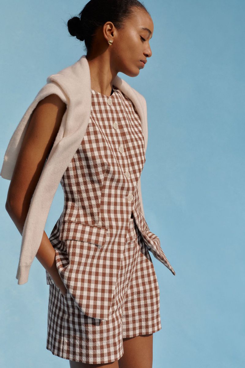 Adult female poses in a brown and white gingham two-piece outfit against a blue backdrop, with a cream sweater draped over her shoulders.