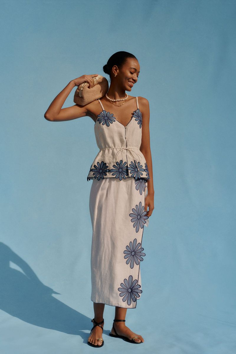 Elegant woman in a floral-patterned top and skirt smiles with a purse over her shoulder against a blue backdrop in a fashion shot.