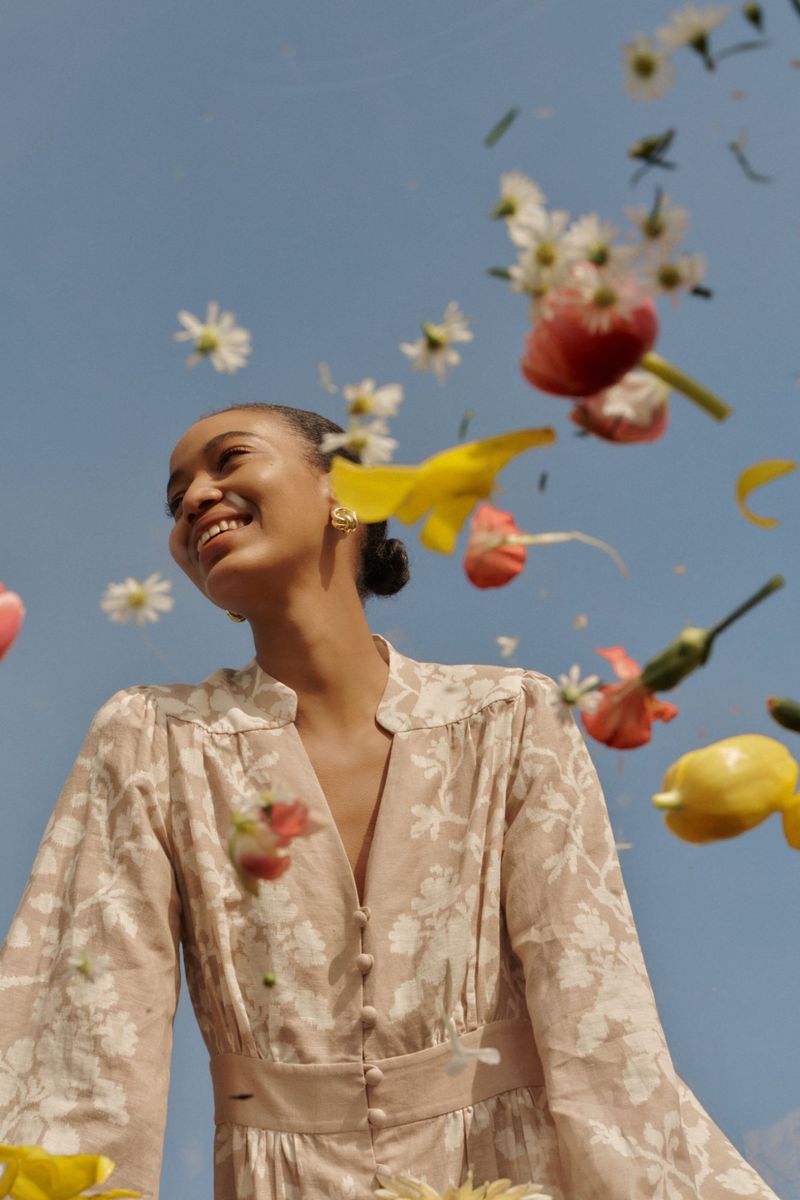 Woman in a floral dress stands under a blue sky with falling flowers, looking up and smiling, creating a serene and fashionable scene.