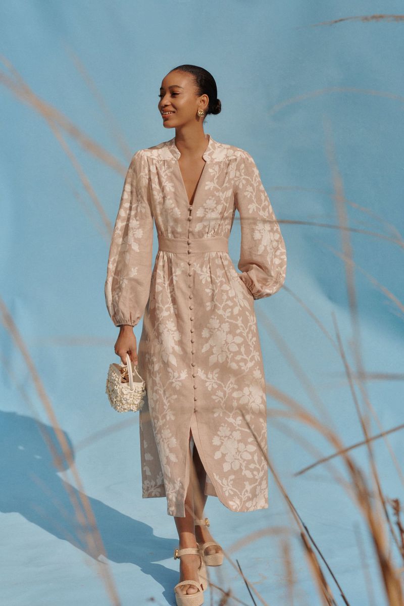 Elegant adult female in long-sleeved floral print dress, holding a beaded handbag, and wearing platform sandals on a blue background.