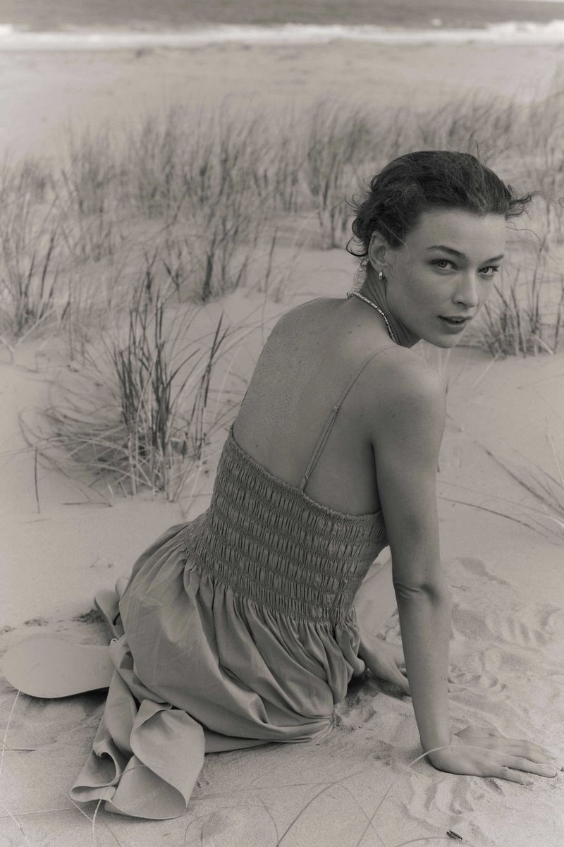 Woman in dress sitting on sandy beach with dunes and ocean in monochrome image, looking at the camera with a slight smile.