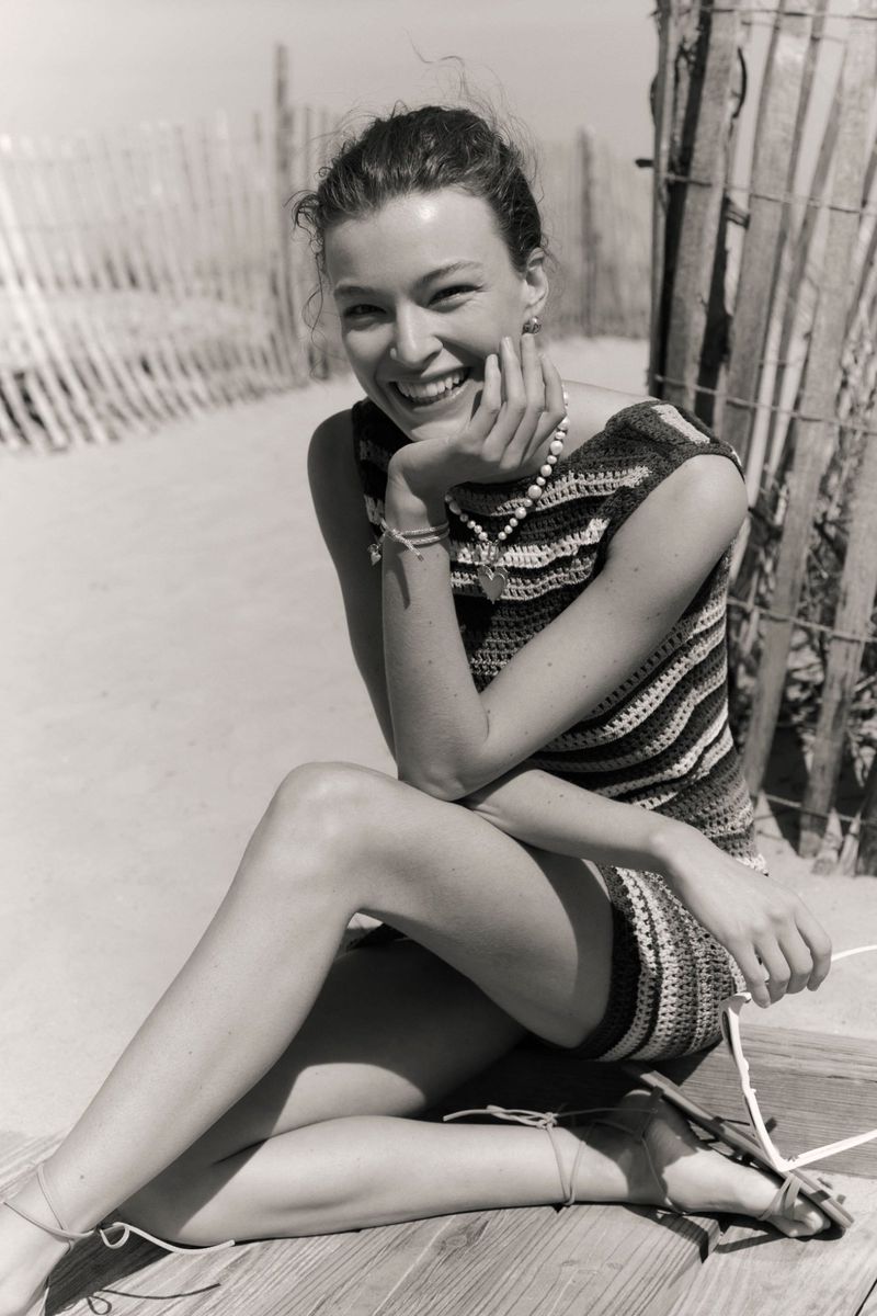 Smiling woman in striped dress poses sitting on boardwalk near sandy beach with wooden fence. Black and white fashion photograph.