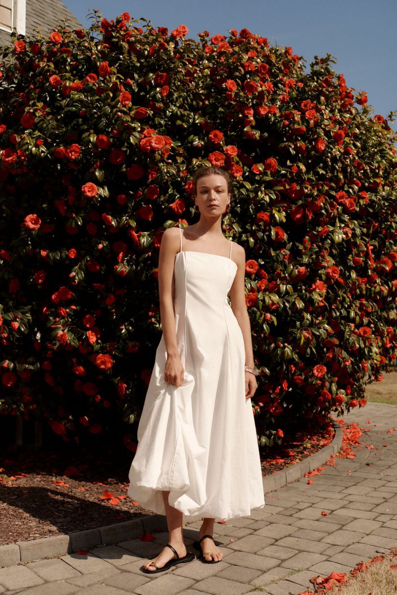 A fashionable woman in a white dress stands in front of a flowering bush with orange blossoms, complemented by a paved stone path.