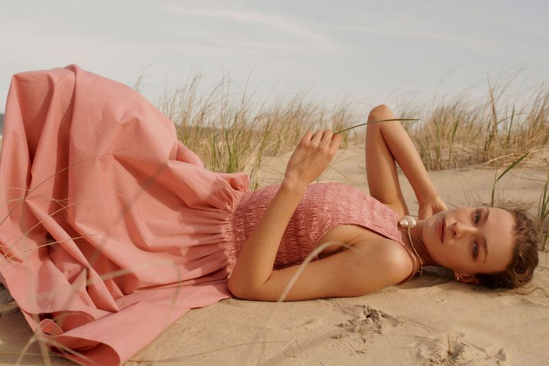 Woman in a pink dress lies on the sand near dune grass, illuminated by soft sunlight, creating a serene and fashionable beach scene.