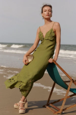 A woman poses in an olive dress with cutouts on a beach, holding a shell purse, wearing platform sandals and a chair in the foreground.