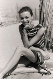 Smiling woman wearing a striped dress and beaded necklace seated outdoors near a wooden fence on a sandy beach.