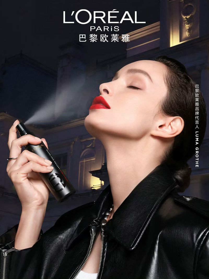 Close-up of an adult female with red lipstick and a black leather jacket, spraying a black bottle of hairspray in an indoor setting.