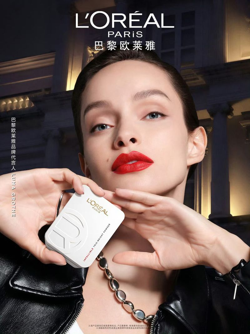Close-up of a woman with red lipstick holding a L'Oréal product, wearing a black leather jacket with city buildings in the background.