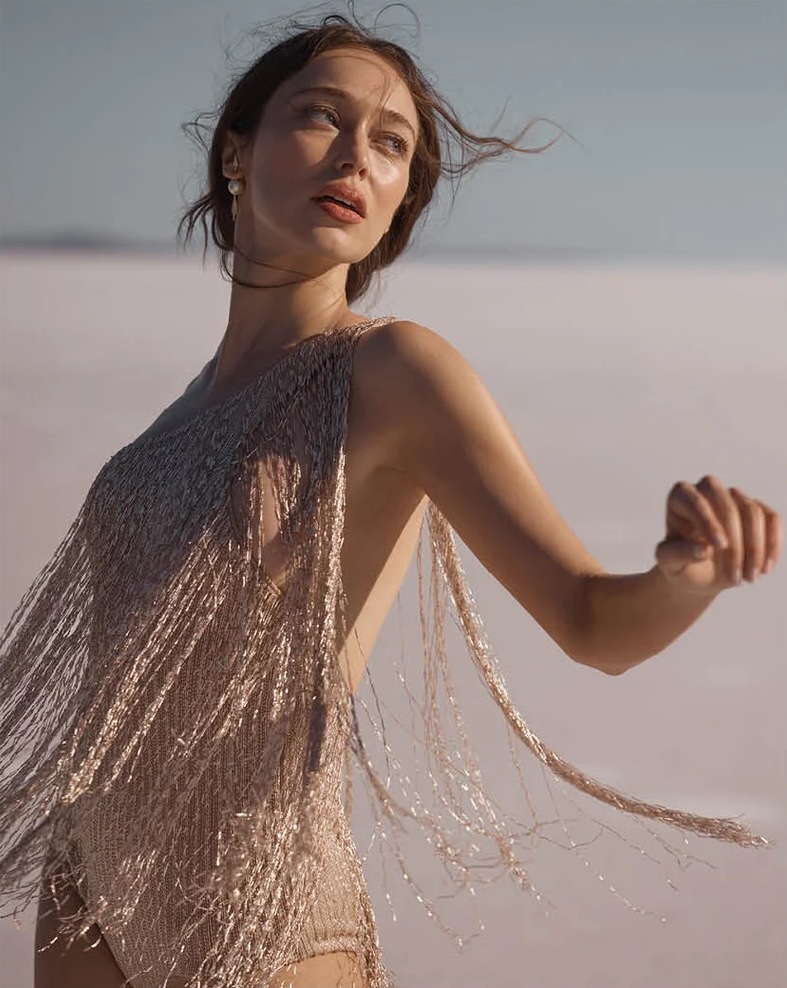 Woman poses on white salt flats wearing a tan fringed bodysuit, with sunlight accentuating her features and the texture of the landscape.
