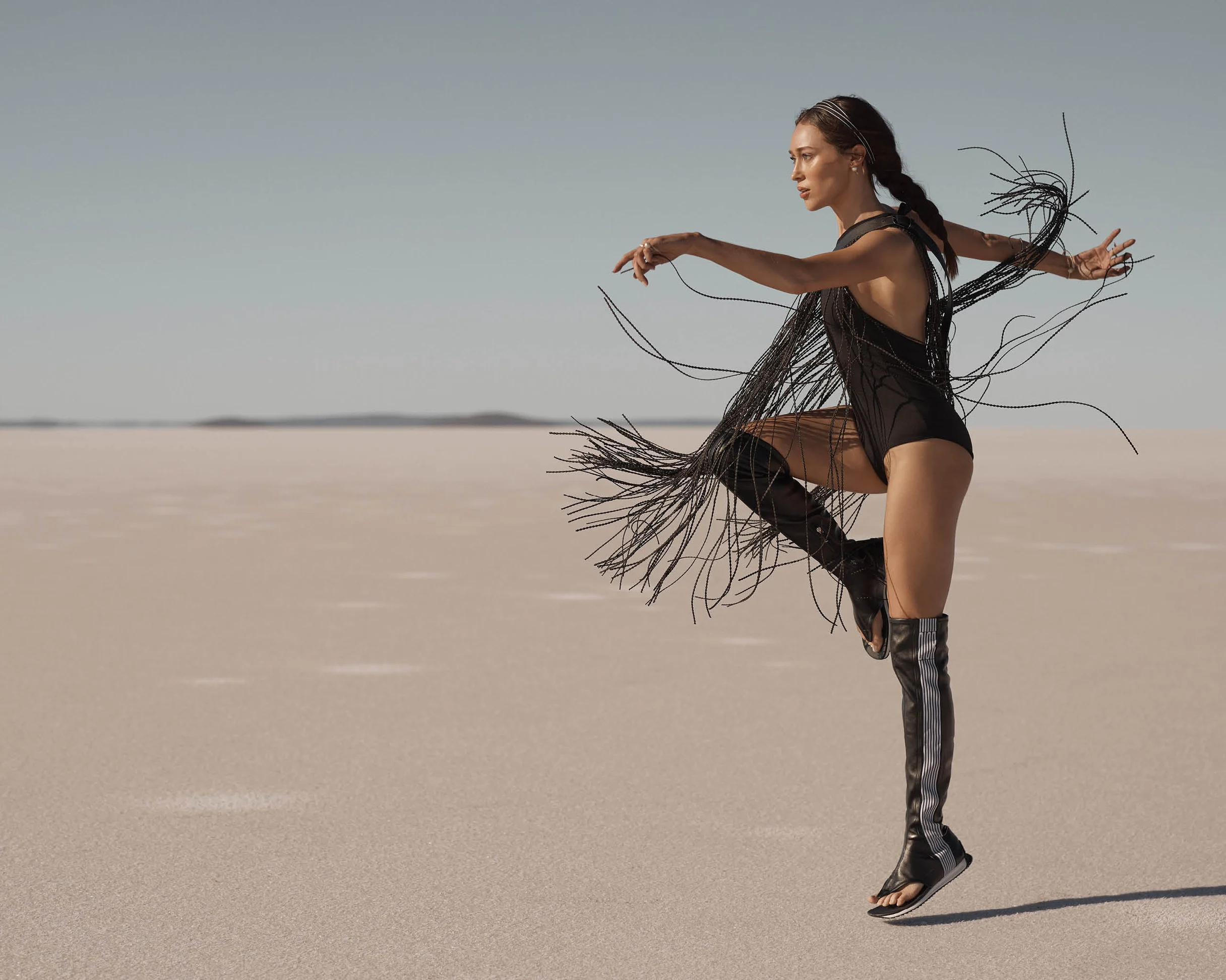 Fashion shot of a woman in a black bodysuit with fringe, posing dynamically in high boots in a desolate, pale landscape under a clear sky.