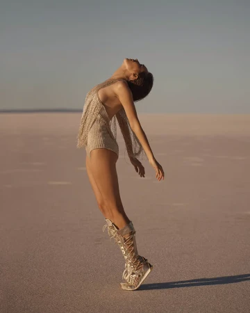 A woman in a glittering bodysuit and boots arches her back on a vast, flat plain under a clear sky, creating a striking silhouette.