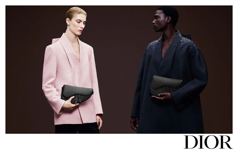 Fashion ad featuring two models, one in pink and the other in navy blue, holding matching black saddle bags, against a brown backdrop.