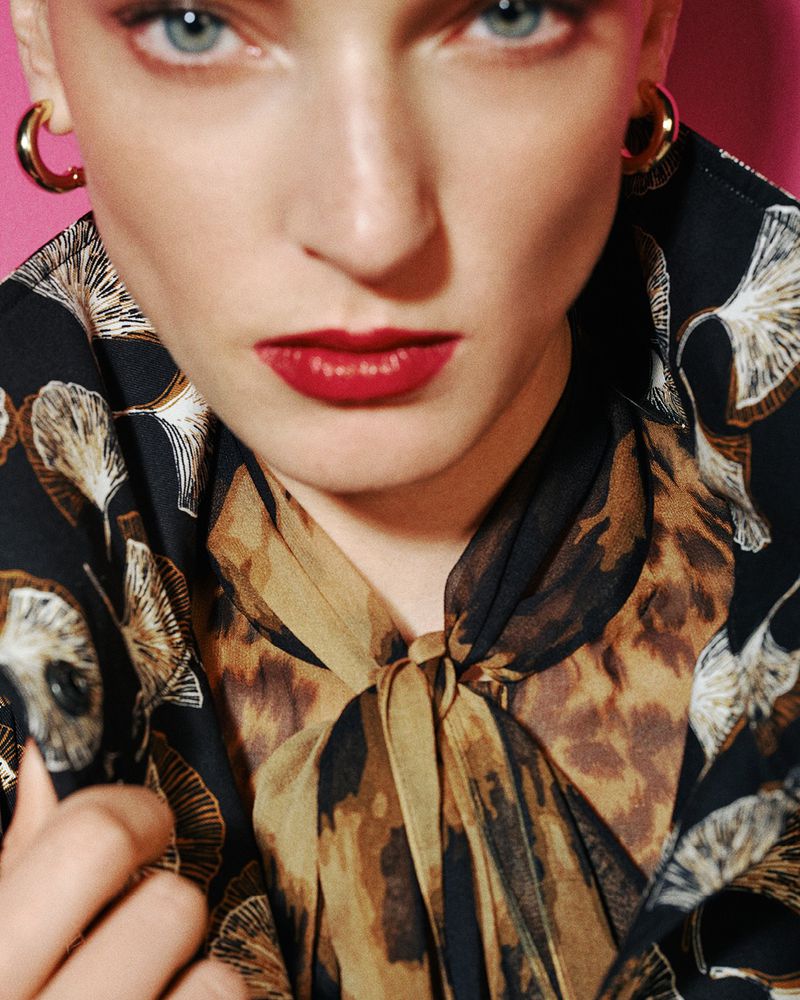 Close-up portrait of a woman with short hair, red lipstick, hoop earrings, and a patterned blouse against a vibrant pink background.