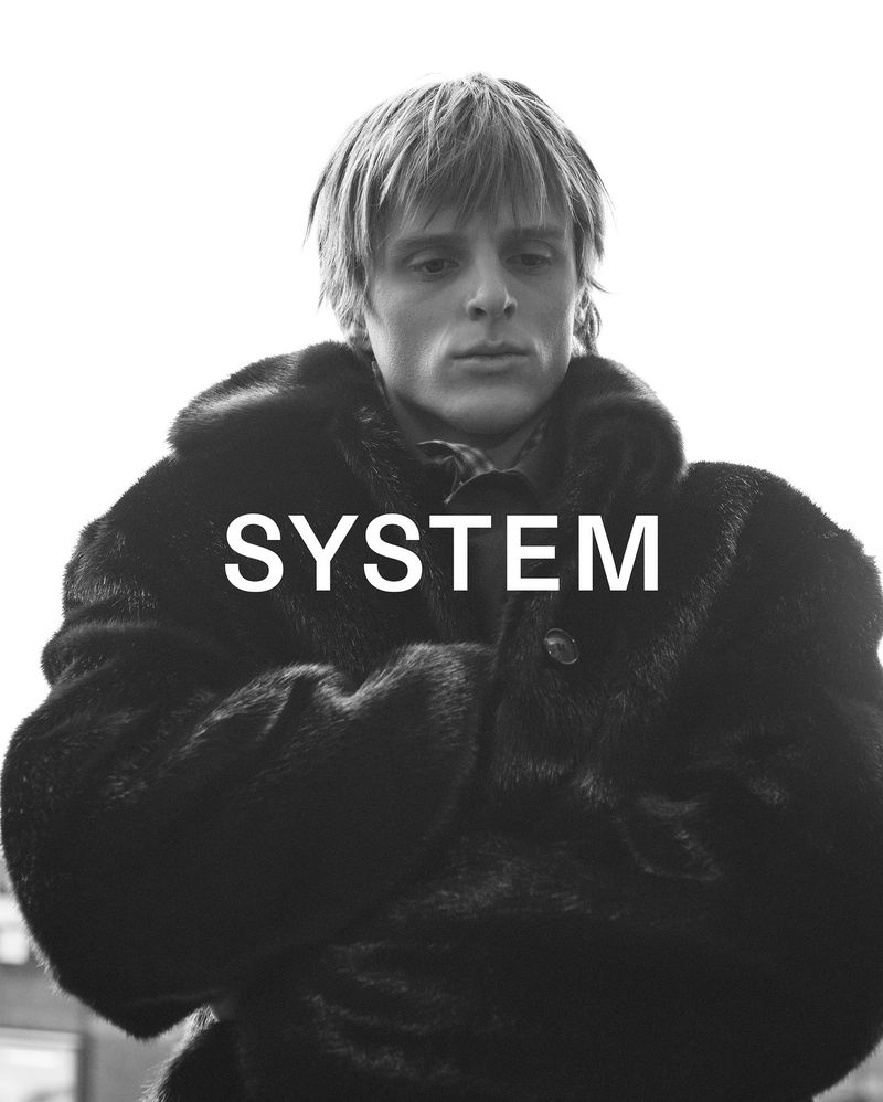 Black and white close-up of a fair-skinned adult male wearing a dark fur coat with the word 'SYSTEM' superimposed on the image.