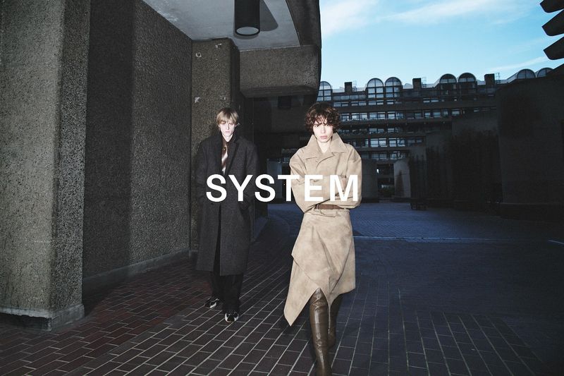 Fashion editorial shot of two adult models wearing a long coat and long dress, posing in an urban architectural setting at dusk.