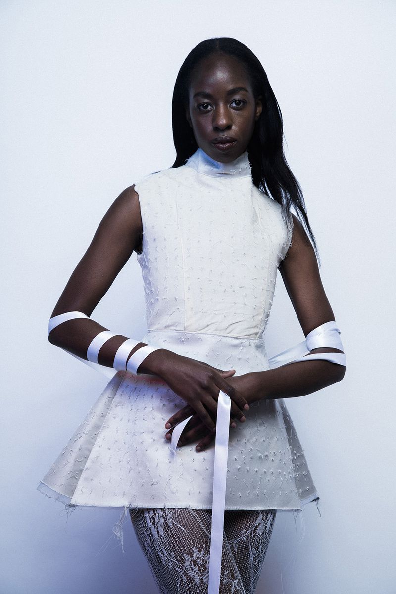 A young Black woman poses in a white, textured mini-dress with ribbon accents, against a pale background in a fashion portrait.