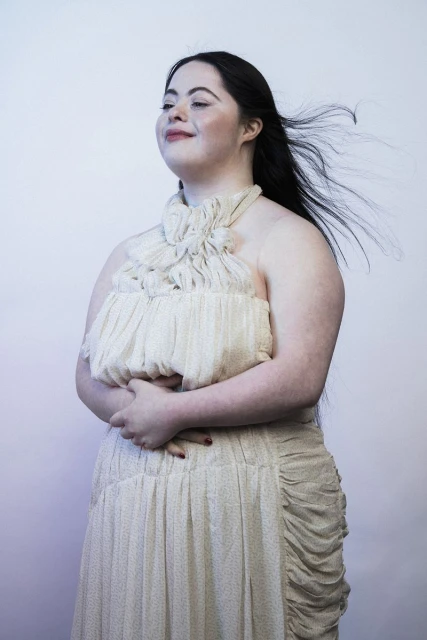 Portrait of a woman with Down syndrome in an evening gown, standing against a soft gradient backdrop, her black hair blowing in the wind.