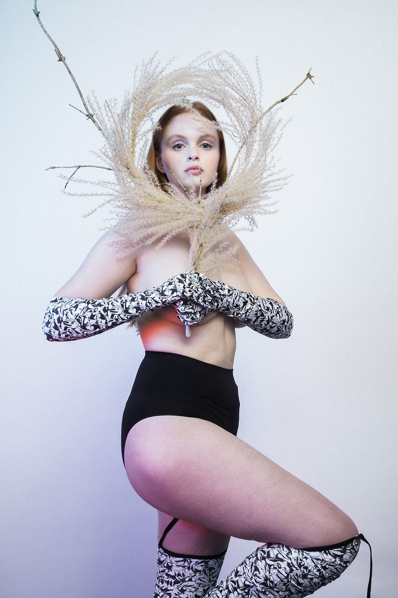Artistic fashion shot of a red-haired model wearing black bottoms and patterned sleeves, holding wheat, standing in a studio.
