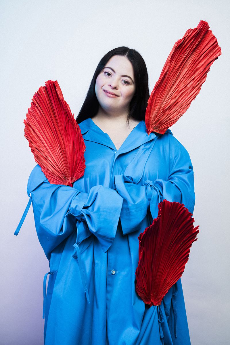 Portrait of a woman with Down syndrome wearing a blue coat and holding large, pleated red fans on a white background.