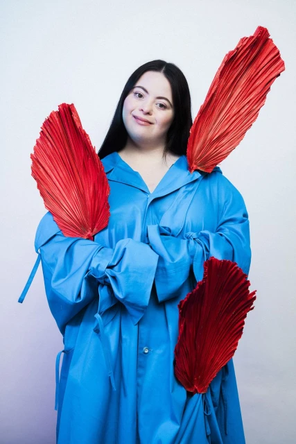 Portrait of a woman with Down syndrome wearing a blue coat and holding large, pleated red fans on a white background.