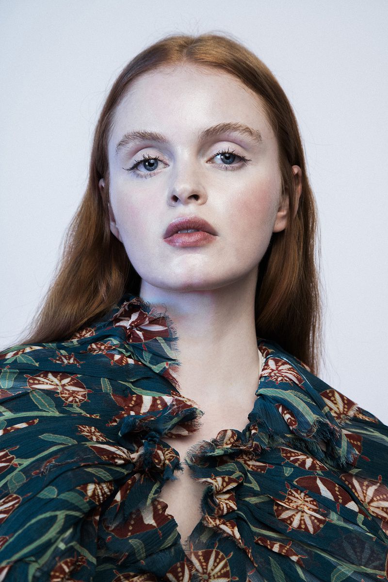 Close-up portrait of a red-haired woman with fair skin, wearing a floral top, against a white background, showcasing beauty and fashion.