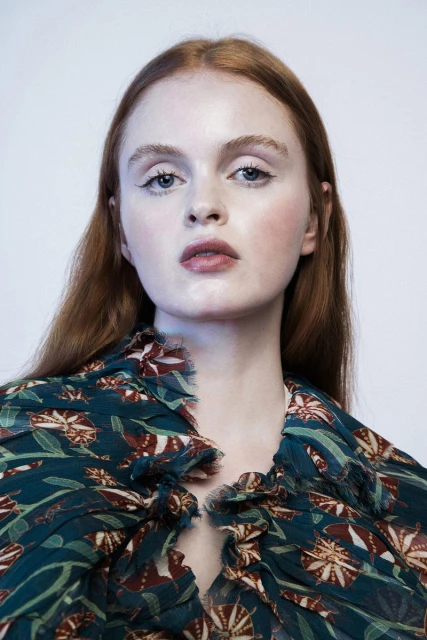 Close-up portrait of a red-haired woman with fair skin, wearing a floral top, against a white background, showcasing beauty and fashion.
