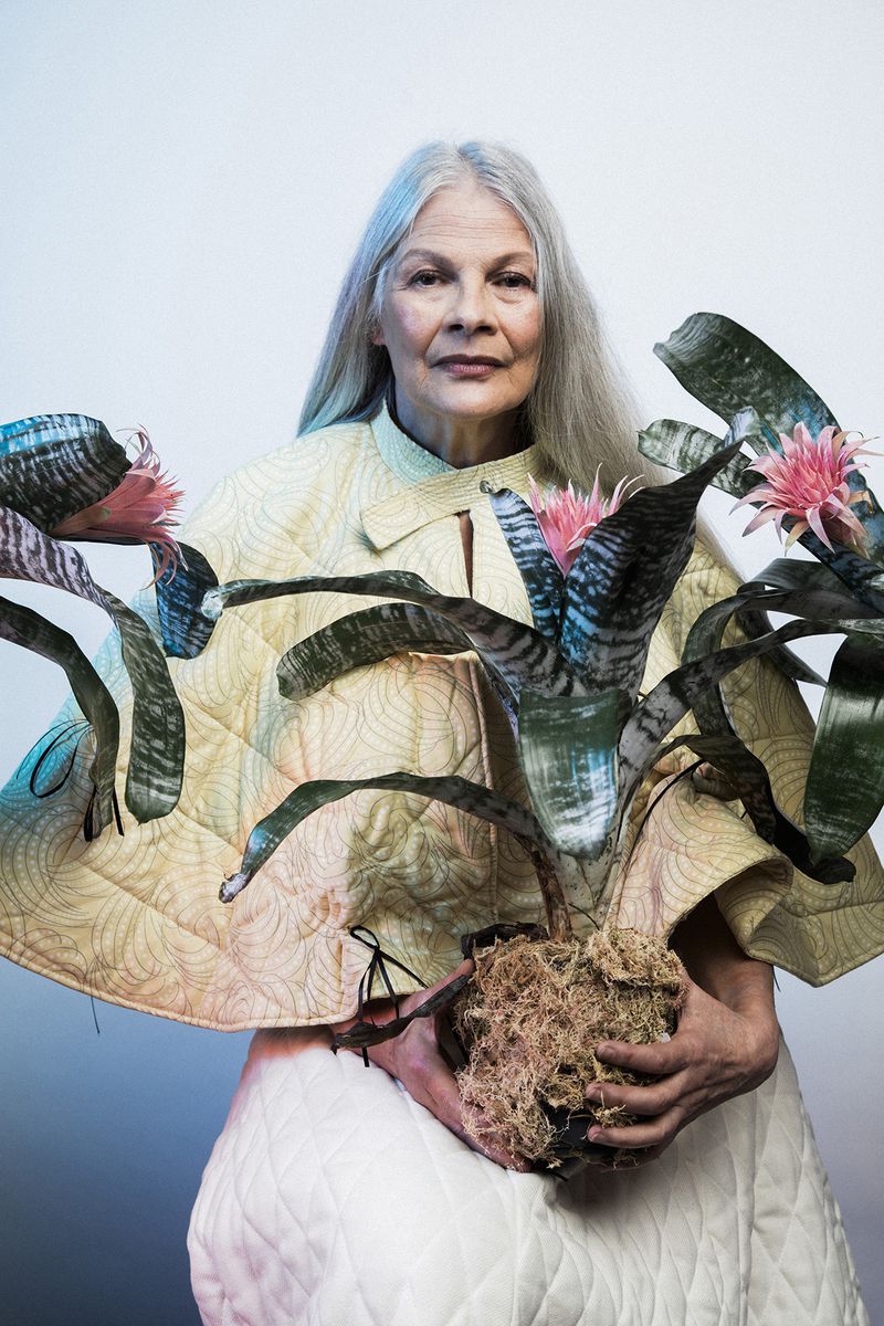 Elderly woman with long grey hair poses indoors wearing a quilted outfit, holding a bromeliad plant in front of a gradient background.