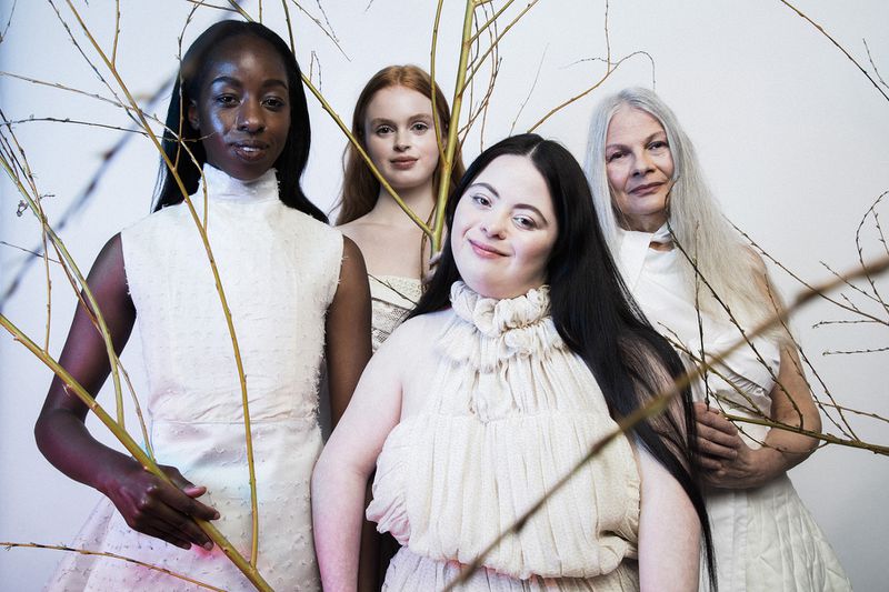 Four diverse women pose in white dresses amid bare branches against a clean backdrop, exuding elegance and a natural aesthetic.